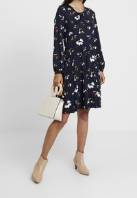 Robe navy avec imprimé floral, manches longues et taille cintrée. Associée à un sac à main crème et des bottines beige.