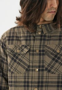 Bruine en zwarte geruite flanellen shirt met een knoopsluiting aan de voorkant, twee borstzakken en metalen knopen. Zachte textuur, standaard kraag, casual pasvorm.