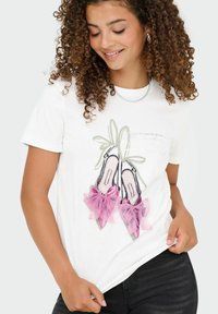 Vrouw met krullend haar, gekleed in een wit shirt met een afbeelding van roze balletschoenen met strikken en een zwarte broek.