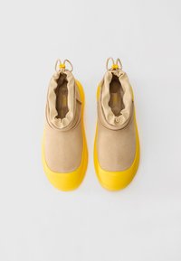 Pantofi slip-on bej cu talpă din cauciuc galbenă și manșete ajustabile cu șnur, prezentați dintr-o perspectiva de sus pe un fundal alb.