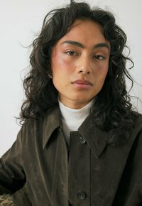 Veste en daim marron avec un col à revers, dotée de deux grands boutons et d'une texture douce. Portée par-dessus un pull col roulé crème. Cheveux bouclés visibles.