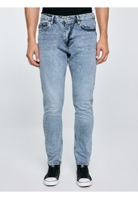 Mees, kes kannab helesiniseid teksapükse ja musti kõrge säärega tossusid, seisab tavalise heleda tausta ees.