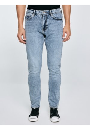 Vīrietis, kurš valkā gaiši zilu džinsa bikses un melnas augstās sporta kurpes, stāv pret vienkrāsainu gaišu fonu.