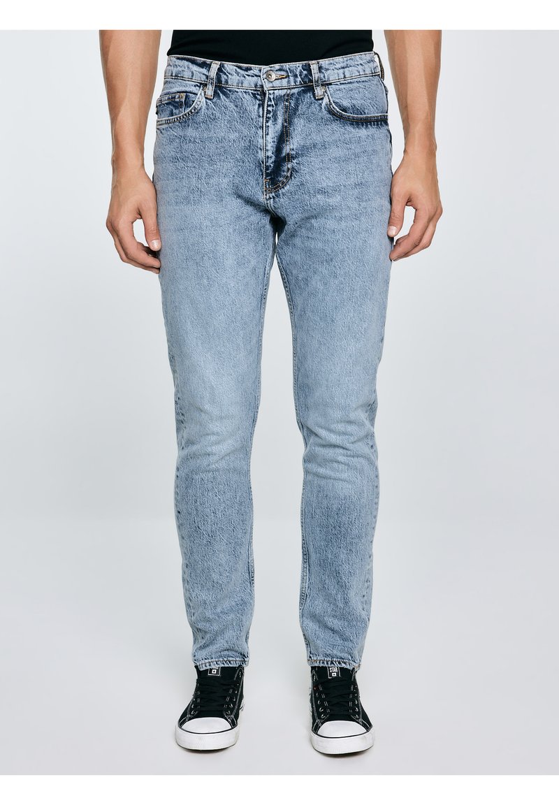 Mees, kes kannab helesiniseid teksapükse ja musti kõrge säärega tossusid, seisab tavalise heleda tausta ees.