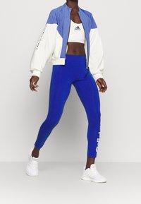 Blå og hvid bomberjakke med stribet design, kombineret med blå leggings og hvide sneakers. Bemærkelsesværdigt logo på sports-bh og leggings.