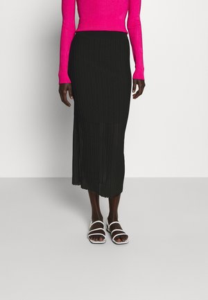 Kobieta w długiej czarnej plisowanej spódnicy, jasnoróżowej bluzce z długim rękawem oraz białych sandałach na paski, stojąca na jednolitym tle.