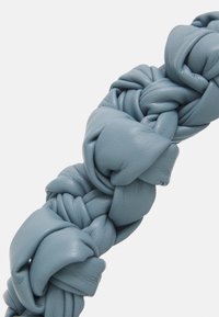 Accessorio in pelle intrecciata di colore azzurro tenue, caratterizzato da una finitura testurizzata con nodi e sezioni lisce, che crea un design stratificato unico.