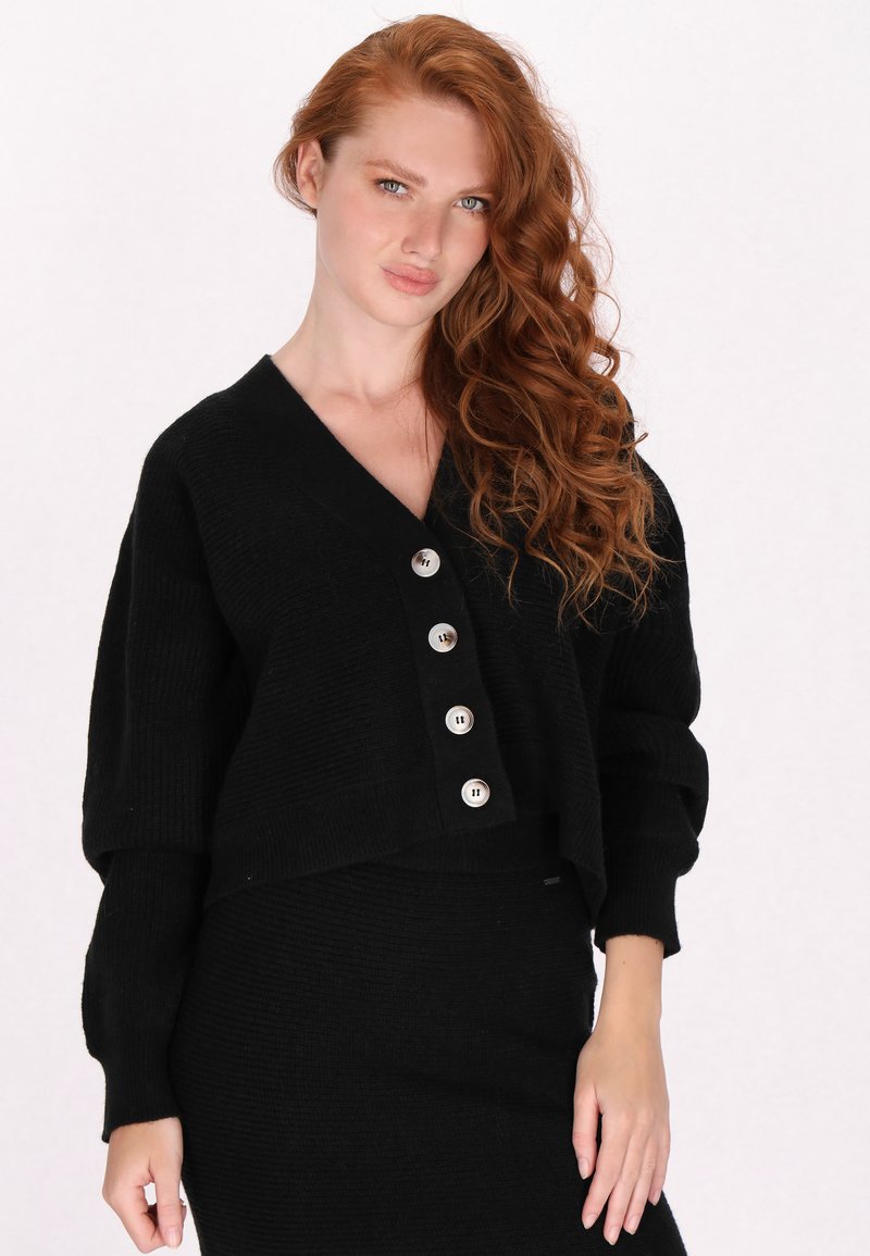 Cardigan nero accorciato con scollo a V, texture a coste e cinque bottoni argentati sul fronte. Maniche lunghe e voluminose. Abbinalo a una gonna.