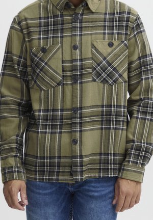 Mand iført olivengrøn og sort ternet skjorte med knapper og to brystlommer samt blå jeans, stående med armene afslappede.