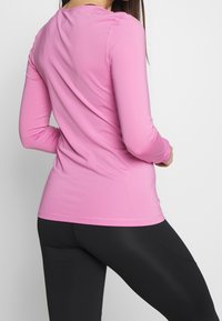 Dlouhý rukáv, přiléhavý top v jasně růžové látce s hladkou texturou. Má jednoduchý kulatý výstřih a mírně zaoblený spodní lem.