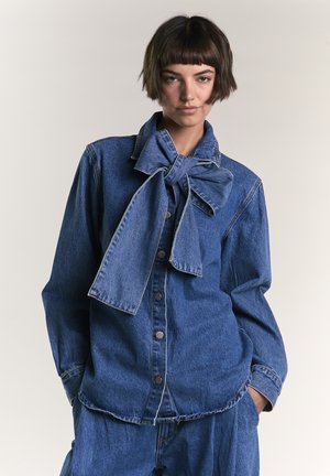 Giovane donna con capelli castani corti che indossa una camicia in denim blu abbottonata con un grande fiocco annodato al colletto e pantaloni in denim coordinati.