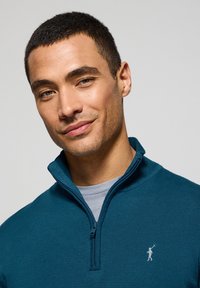 Teal zip-up tröja i texturerat material med hög krage. Har en liten ljusgrå logotyp på vänster bröst.