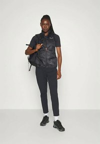 Schwarze Puffer-Weste über einem kurzärmligen schwarzen Oberteil, kombiniert mit schmalen schwarzen Hosen und schwarzen Sneakers. Die Weste verfügt über einen Reißverschluss und Taschen.