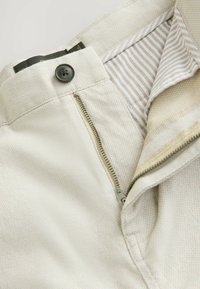 Pantaloni de culoare deschisă dintr-un material texturat, cu închidere prin fermoar și nasture, și căptușeală în dungi pe interiorul taliei.