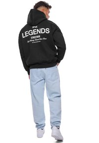 Sort hoodie med teksten "LEGENDS STREETWEAR" i hvid på ryggen, parret med lyseblå jeans og hvide sneakers. Casual streetwear stil.