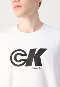T-shirt in cotone bianco con un grande logo "CK" nero con dettaglio circolare. Include il nome del marchio "Calvin Klein" in caratteri più piccoli sotto.