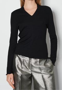 Svart långärmad topp med V-ringning, med en något skrynklig textur och ojämn nederkant. Matchad med glänsande metalliska silverbyxor.