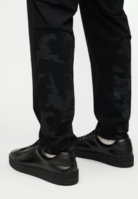 Μαύροι αθλητικοί παντελόνες με υφασμάτινες καμουφλάζ λεπτομέρειες στα πόδια, σε συνδυασμό με μαύρα δερμάτινα αθλητικά παπούτσια με λεία υφή και μινιμαλιστικό σχεδιασμό.