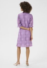 Violeta mežģīņu kleita ar pūkām pie piedurknēm, rotāta ar ziedu rakstiem un brīvu piegriezumu, sasniedz vidusceli. Modelis valkā bežīgas slingback kurpes.