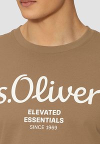 Hnědé tričko s kulatým výstřihem, které má bílé potisk, jež zní "s.Oliver ELEVATED ESSENTIALS SINCE 1969." Hladká textura, volný střih.