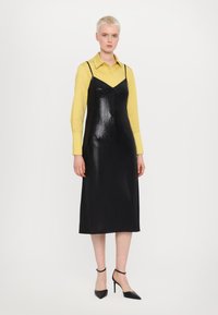 Schwarzes Midikleid mit glänzender Textur und verstellbaren Spaghettiträgern, getragen über einem gelben Shirt. Kombiniert mit schwarzen spitzen Pumps.