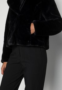 Veste en fausse fourrure noire avec un large col, devant ouvert et manches longues. Comprend une poche latérale et une finition douce et texturée.