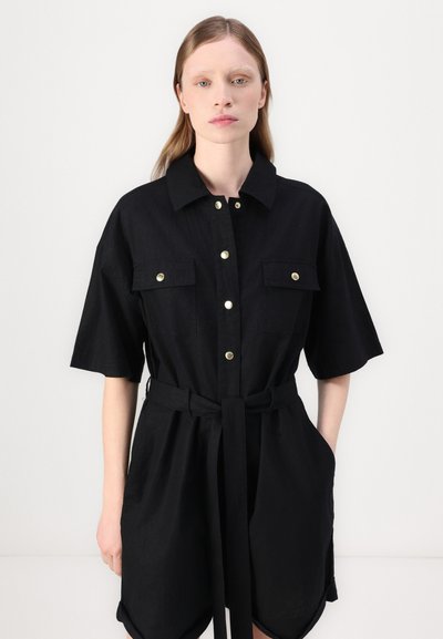 Robe noire à manches courtes, devant boutonné, deux poches poitrine, taille ceinturée, fabriquée en tissu lisse et dotée de accents en boutons dorés.