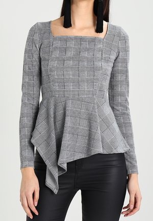 Nainen, joka pukeutunut harmaaseen ruudulliseen pitkähihainen peplumt- paitaan, jossa on epäsymmetrinen helma ja mustat tupsukorvakorut, yhdistettynä mustiin housuihin.