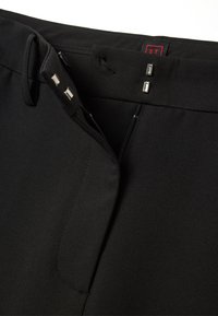 Pantalons noirs avec une texture lisse, fermeture éclair et deux accents métalliques à la taille. Comprend une étiquette rouge avec "ST."
