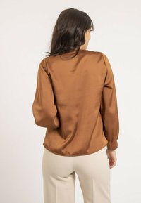 Chemise en satin marron à manches longues, avec un ourlet arrondi et un éclat subtil. Présente une coupe décontractée et des détails de couture minimalistes au niveau des poignets.