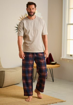 Beige short-sleeve t-shirt paired with blue and red plaid flannel pants, featuring a loose fit and soft texture. Model stands barefoot.