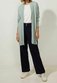 Ljus mintgrön cardigan över en vit topp, i kombination med marinblå vida byxor. Vita sneakers med en strukturerad design.