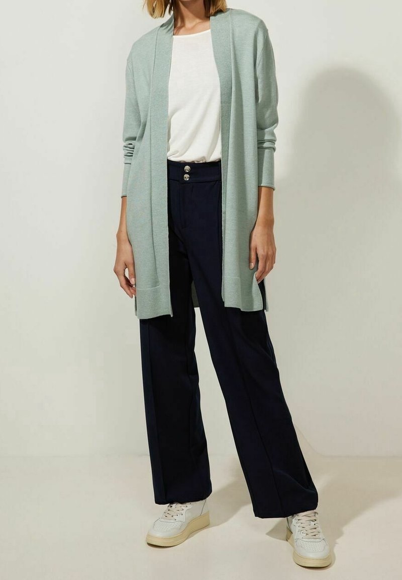 Ljus mintgrön cardigan över en vit topp, i kombination med marinblå vida byxor. Vita sneakers med en strukturerad design.