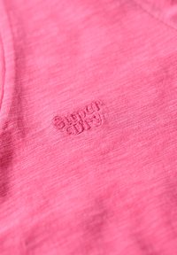 Camiseta rosa de tela suave y texturizada. Presenta un logotipo bordado en rosa a juego, situado en el centro de la tela.