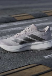 Hellgrauer Sportschuh mit schwarzen Streifen auf Asphalt in der Nähe von gelb-schwarzen Bodenschwellen und einem Zebrastreifen.