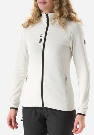 Woman wearing a white zipped jacket with black zipper and logo, paired with black pants, standing against a plain white background.