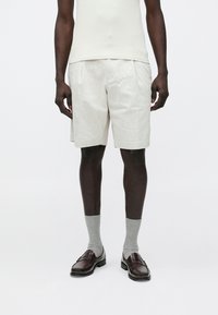 Shorts en lin beige avec plis, associés à des chaussettes grises et des mocassins en cuir marron. Le mannequin porte un débardeur crème ajusté.