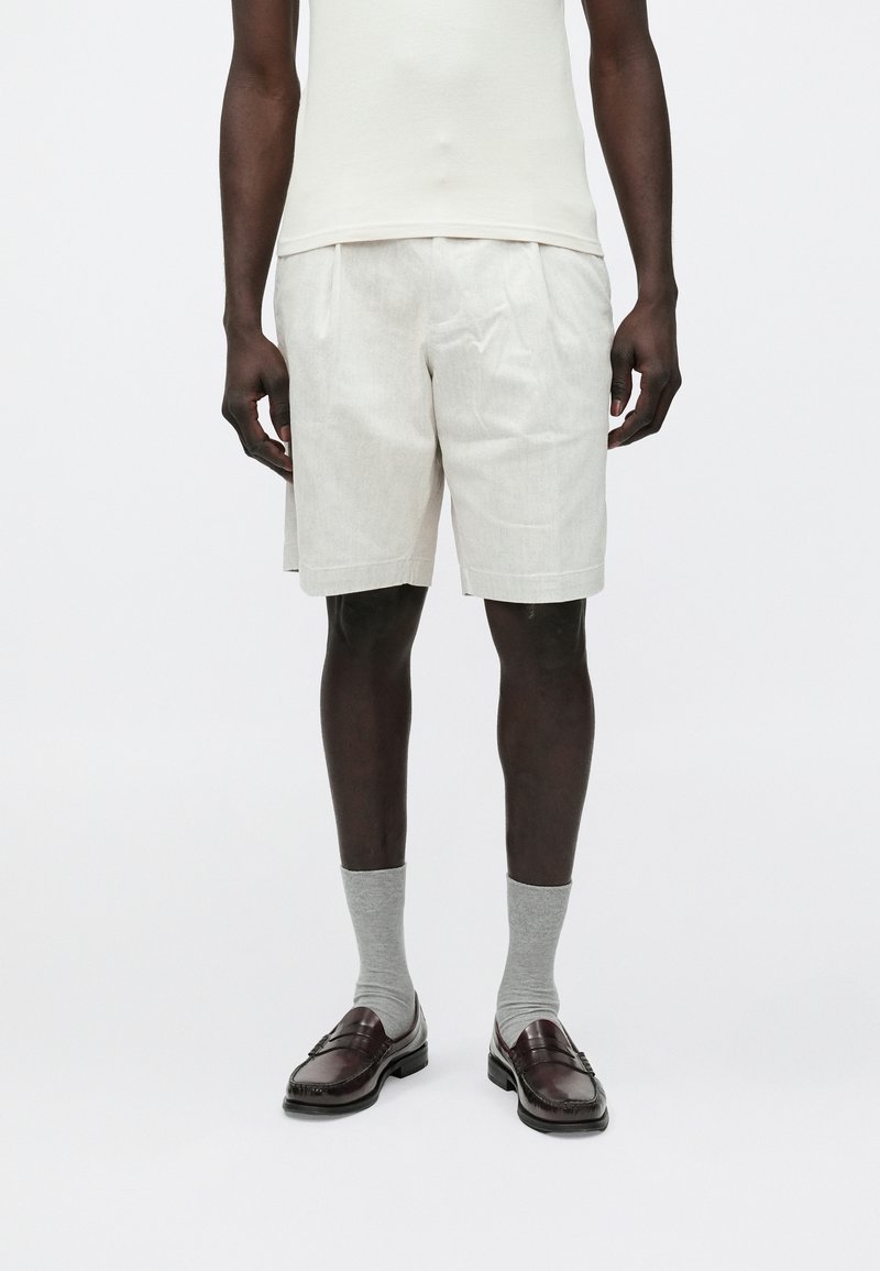 Shorts en lin beige avec plis, associés à des chaussettes grises et des mocassins en cuir marron. Le mannequin porte un débardeur crème ajusté.