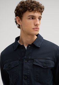 Giacca di jeans blu navy con due tasche sul petto e bottoni neri. Presenta un colletto classico e una trama testurizzata, progettata per una vestibilità casual.