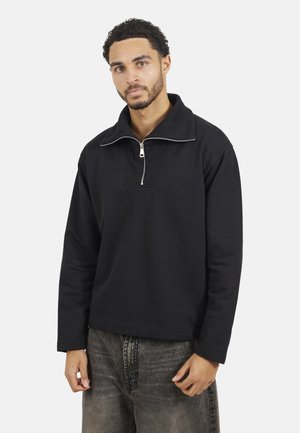 Felpa nera con colletto a mezza zip, texture a coste e un sottile dettaglio sul pannello frontale; vestibilità rilassata, abbinata a jeans sbiaditi.