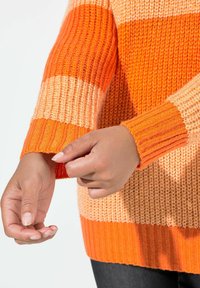 Sweter dziany w pomarańczowe i brzoskwiniowe poziome paski; ściągacze na mankietach i u dołu; lekki, teksturowany materiał. Ręce poprawiające rękaw.