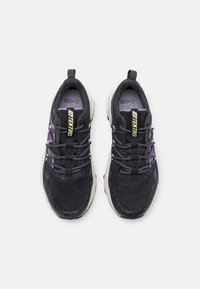 Schwarze Sportschuhe aus strukturiertem Material, mit lila Akzenten und gemusterten Schnürsenkeln. Sie verfügen über eine dicke weiße Sohle und eine Zuglasche an der Rückseite.