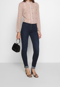 Light pink, polka-dot blouse with long sleeves and button front, paired with dark skinny jeans and black handbag. Heeled sandals complete the look.