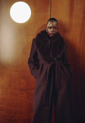 Manteau violet foncé avec un large col en fourrure, cintré à la taille, doté de manches longues et de profondes poches, situé devant un fond en bois.