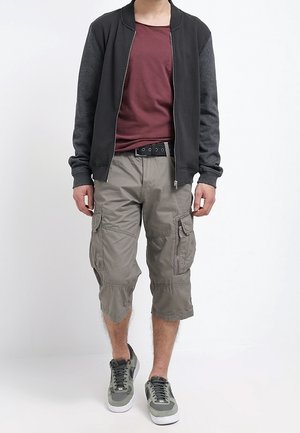 Mann trägt eine schwarze Zip-Up-Jacke, ein kastanienbraunes T-Shirt, graue Cargo-Shorts und graue Turnschuhe, steht vor einem schlichten weißen Hintergrund.