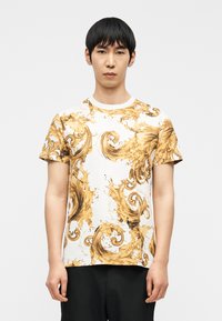 Νεαρός με κοντά μαύρα μαλλιά που φοράει λευκό μπλουζάκι τυπωμένο με χρυσό κίτρινο στροβιλιζόμενο μπαρόκ μοτίβο και μαύρο παντελόνι.