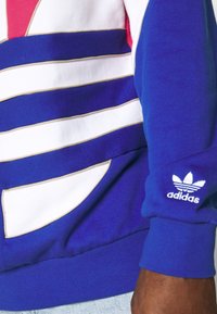 Blå och vit sweatshirt med horisontella ränder, med guldaccenter och en sidoficka, samt en broderad Adidas-logotyp på ärmen.