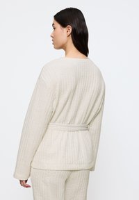 Robe trapuntato beige chiaro con maniche lunghe, vita con cintura e tessuto testurizzato. Il design presenta sottili linee verticali e una vestibilità rilassata.