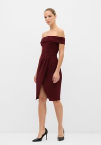Burgundi off-the-shoulder mekko, jossa on tiukka vartalo, roikkuva yksityiskohta ja edessä halkio. Sileä pinta yhdistettynä teräväkärkisiin mustiin korkokenkiin.
