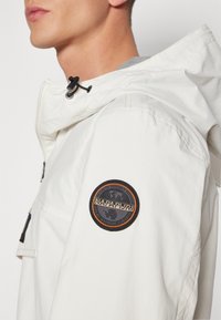 White lightweight jacket with a fitted hood. Features a black cord adjuster and a round, multicolor patch on the left sleeve.